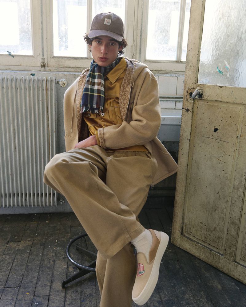 Model Max Fauviau seated on a stool wearing a camel hooded coat, plaid scarf, beige trousers, and baseball cap.