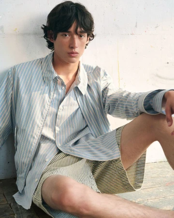 Model wearing a blue striped button-down shirt and shorts seated against a textured white wall.