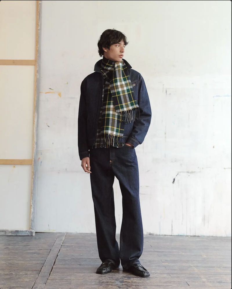 Model wearing a dark denim jacket, matching wide-leg jeans, and a green plaid scarf against a distressed white studio wall.