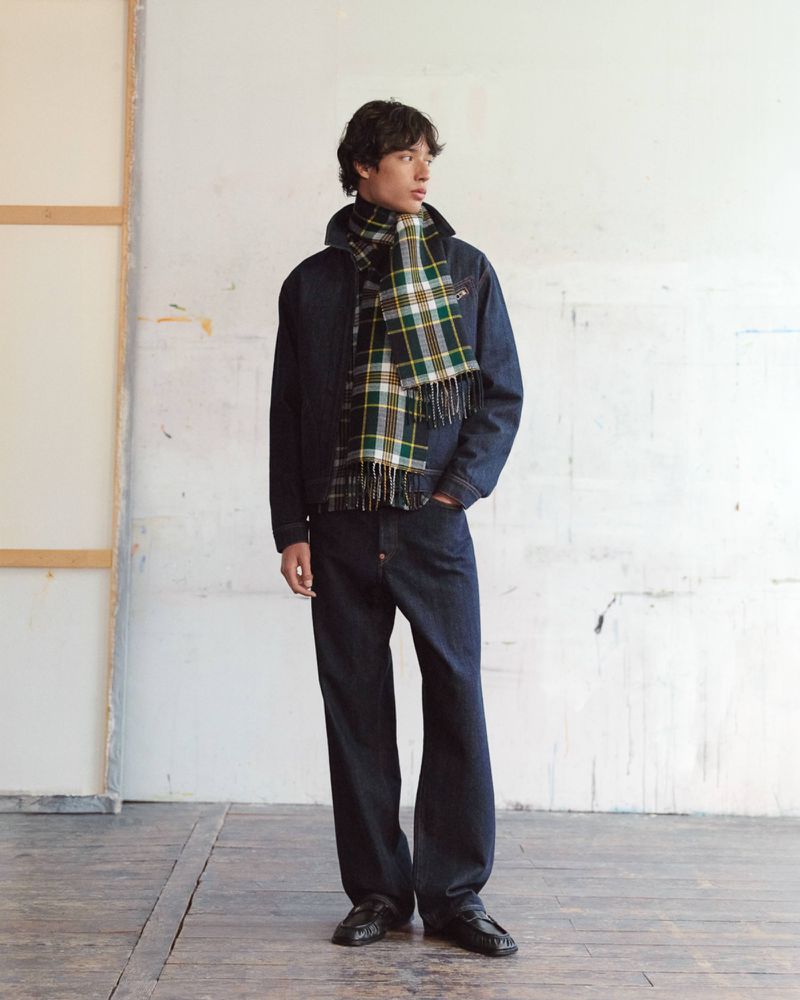 Model wearing a dark denim jacket and plaid scarf standing against an off-white textured studio wall for Kenzo Pre-Spring 2027.