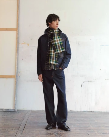 Model wearing a dark denim jacket and plaid scarf standing against an off-white textured studio wall for Kenzo Pre-Spring 2027.
