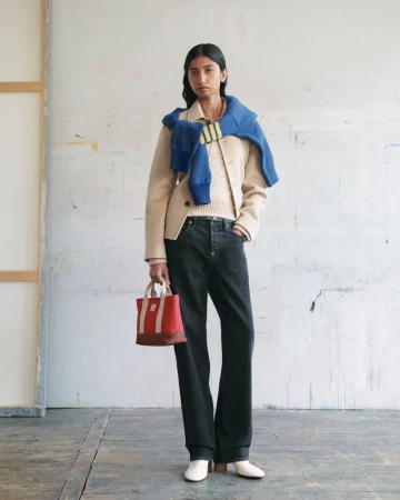 Model standing against a worn wall wearing a beige jacket, dark jeans, and a blue sweater draped over shoulders while holding a red bag