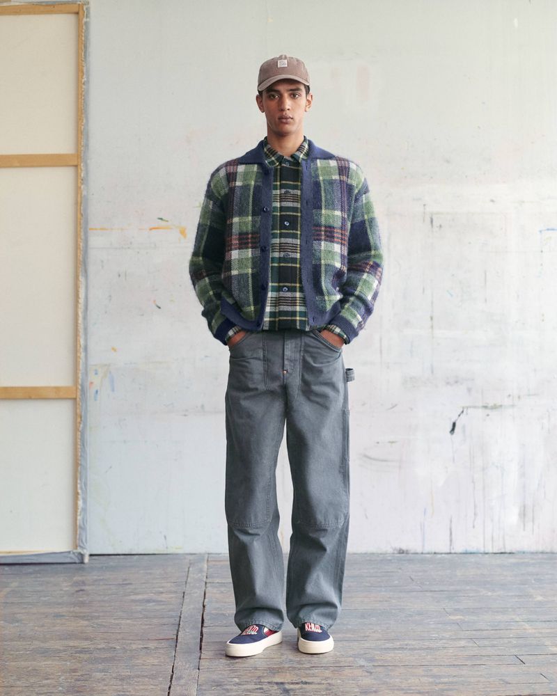 Model wearing a green and navy plaid cardigan over a matching shirt with loose grey trousers and a baseball cap.