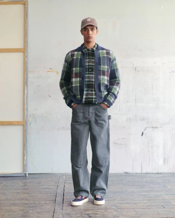 Model wearing a green and navy plaid cardigan over a matching shirt with loose grey trousers and a baseball cap.