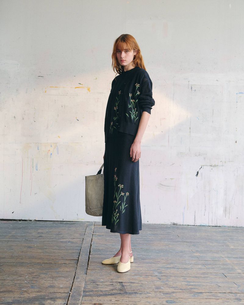 Model wearing a black floral embroidered top and matching midi skirt while holding a patterned tote bag on wooden flooring.
