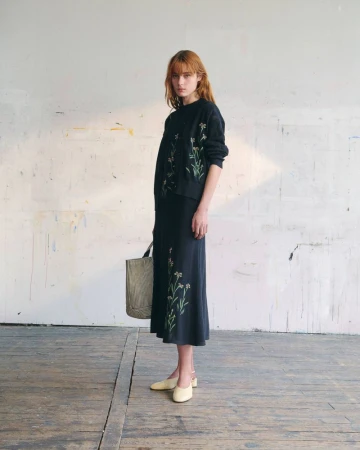 Model wearing a black floral embroidered top and matching midi skirt while holding a patterned tote bag on wooden flooring.