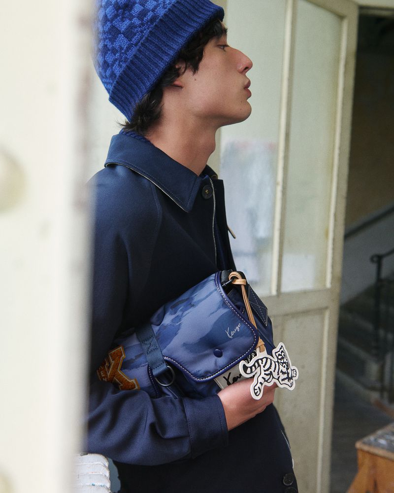 Young man in profile wearing a blue knit beanie, navy jacket, and a blue crossbody bag with a tiger charm by a doorway.