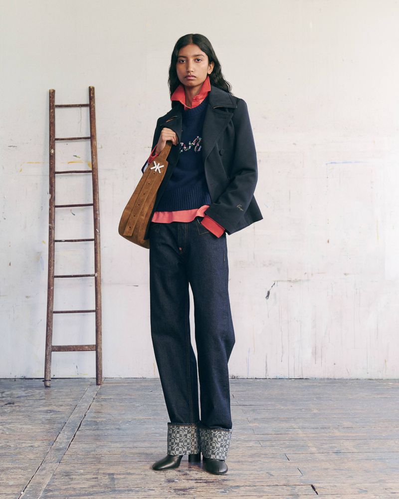 Model wearing a dark jacket, navy sweater, wide jeans with patterned cuffs, and a brown suede bag next to a wooden ladder.
