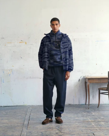 Model standing against a worn studio wall wearing a blue patterned fleece jacket and dark trousers for Kenzo Pre-Spring 2027.