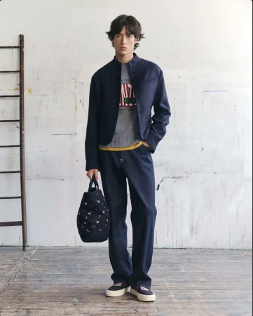 Model wearing a dark blue jacket and trousers holding a patterned tote bag against a weathered wall background