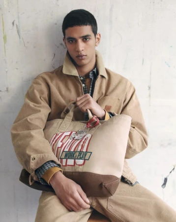 Model seated in a tan canvas jacket and trousers holding a large Kenzo tote bag against a textured studio backdrop.