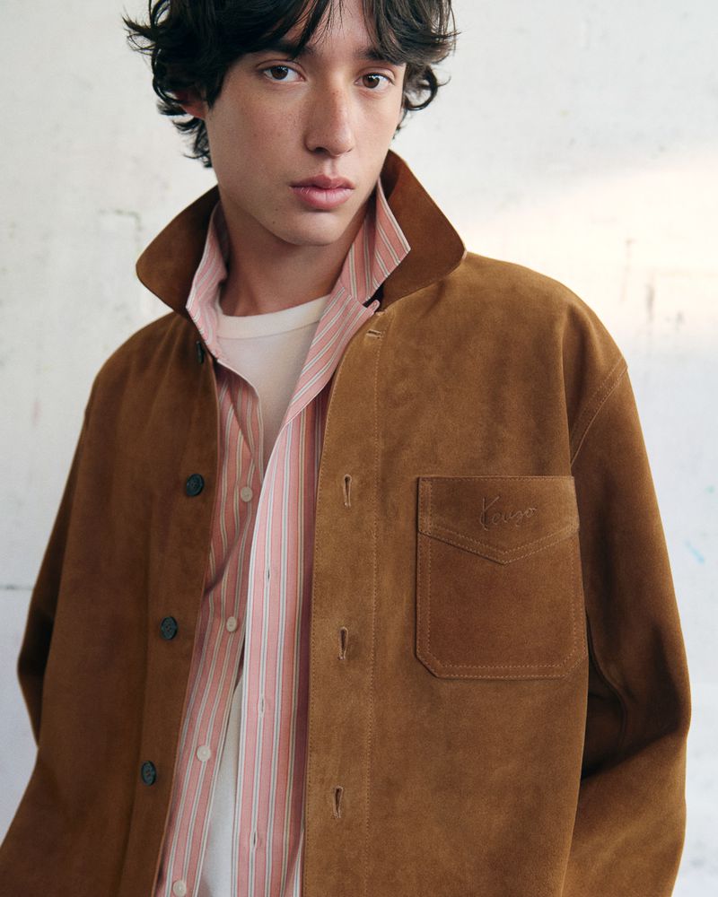 Close-up portrait of a male model wearing a brown suede jacket over a striped shirt against a plain wall.