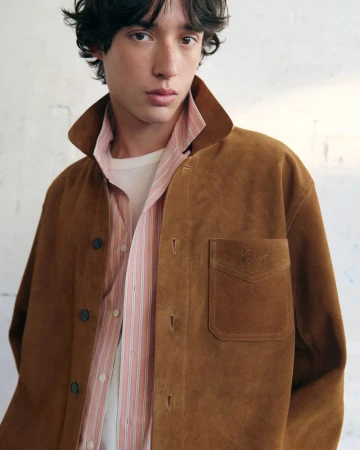 Close-up portrait of a male model wearing a brown suede jacket over a striped shirt against a plain wall.