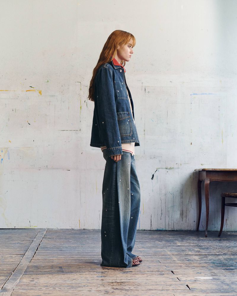 Model standing in profile wearing a speckled denim jacket and extra-wide denim trousers against a studio wall.
