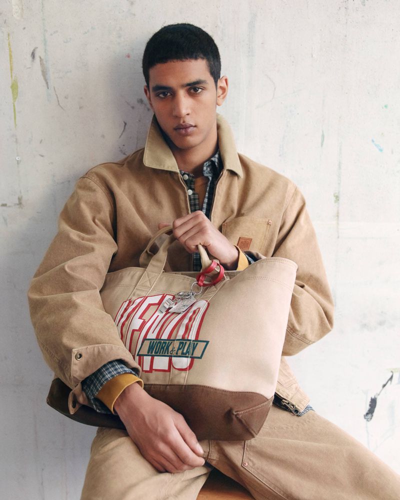 Model seated against a mottled wall wearing a tan canvas jacket and holding a branded tote bag.