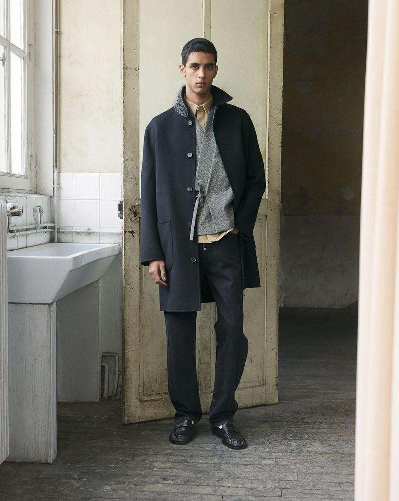 Full-length portrait of a male model wearing a dark coat and trousers standing in an interior space near a doorway.