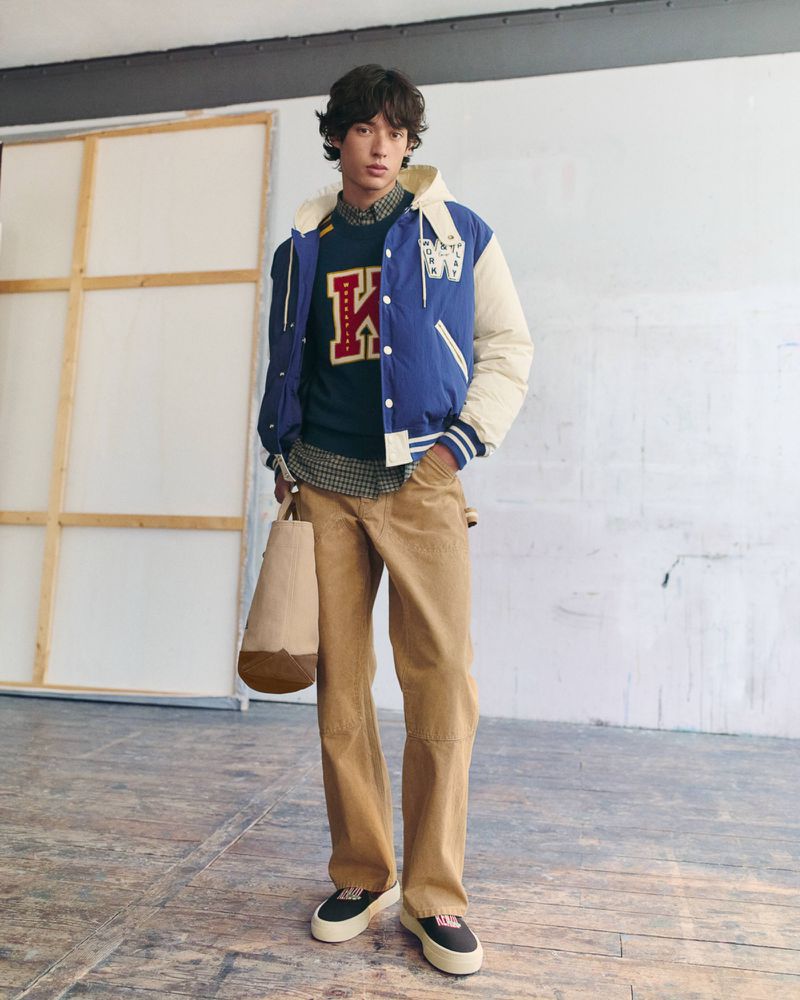 Model wearing a blue Kenzo varsity jacket, green K-logo sweater, tan trousers, and holding a canvas tote bag.