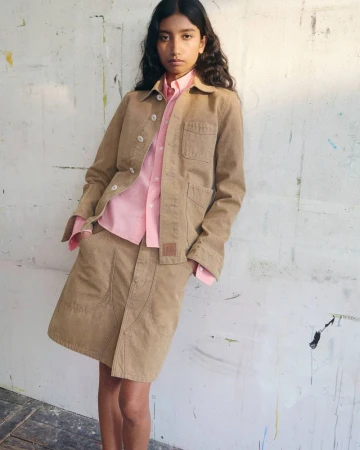 Model wearing a tan jacket and skirt over a pink shirt posing against a textured white wall for Kenzo Pre-Spring 2027.