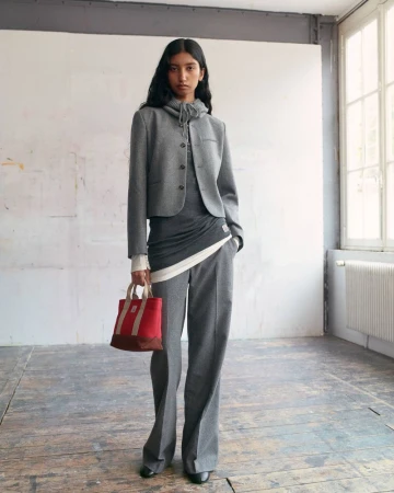 Model standing in a grey jacket and trousers holding a small red handbag in an interior room