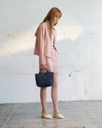 Model with long red hair wearing a pink striped jacket and shorts holding a blue handbag against a white studio wall