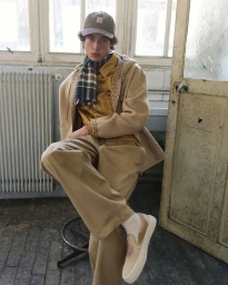 Model Max Fauviau seated on a stool wearing a camel hooded coat, plaid scarf, beige trousers, and baseball cap.
