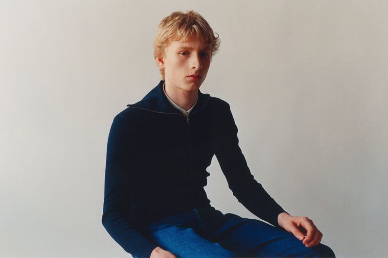 Young male model with blonde hair wearing a dark blue zip-up sweater sitting against a neutral off-white background.