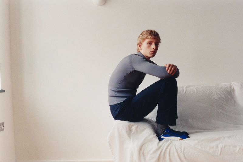 A model in a grey mock-neck sweater and navy trousers sits perched on a white sheet-covered sofa against a plain wall.