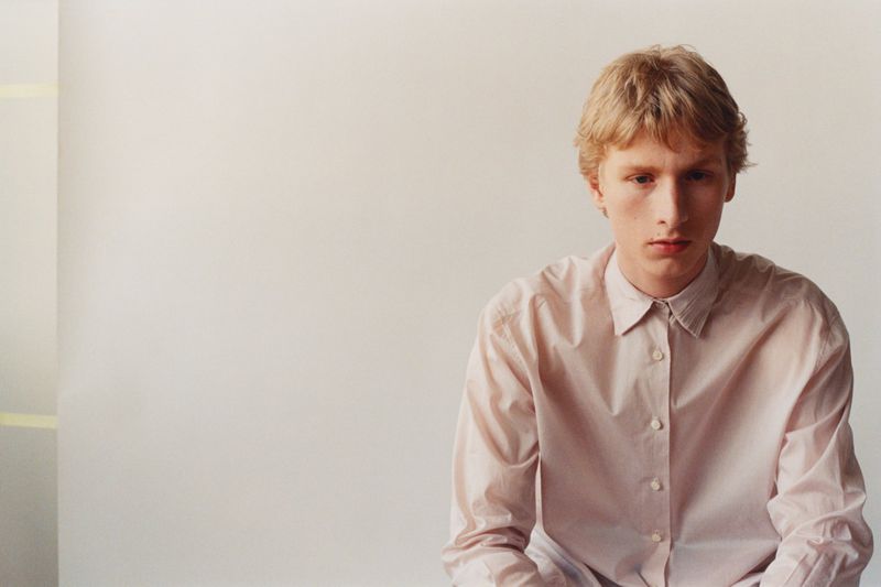 Blonde man wearing a pale pink button-down collared shirt seated against a light studio backdrop.