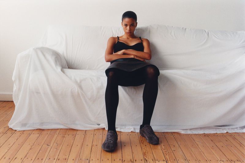 Model Honey Ordonez seated with arms crossed on a white sheet-covered sofa over wooden floors for Puma x Jil Sander.