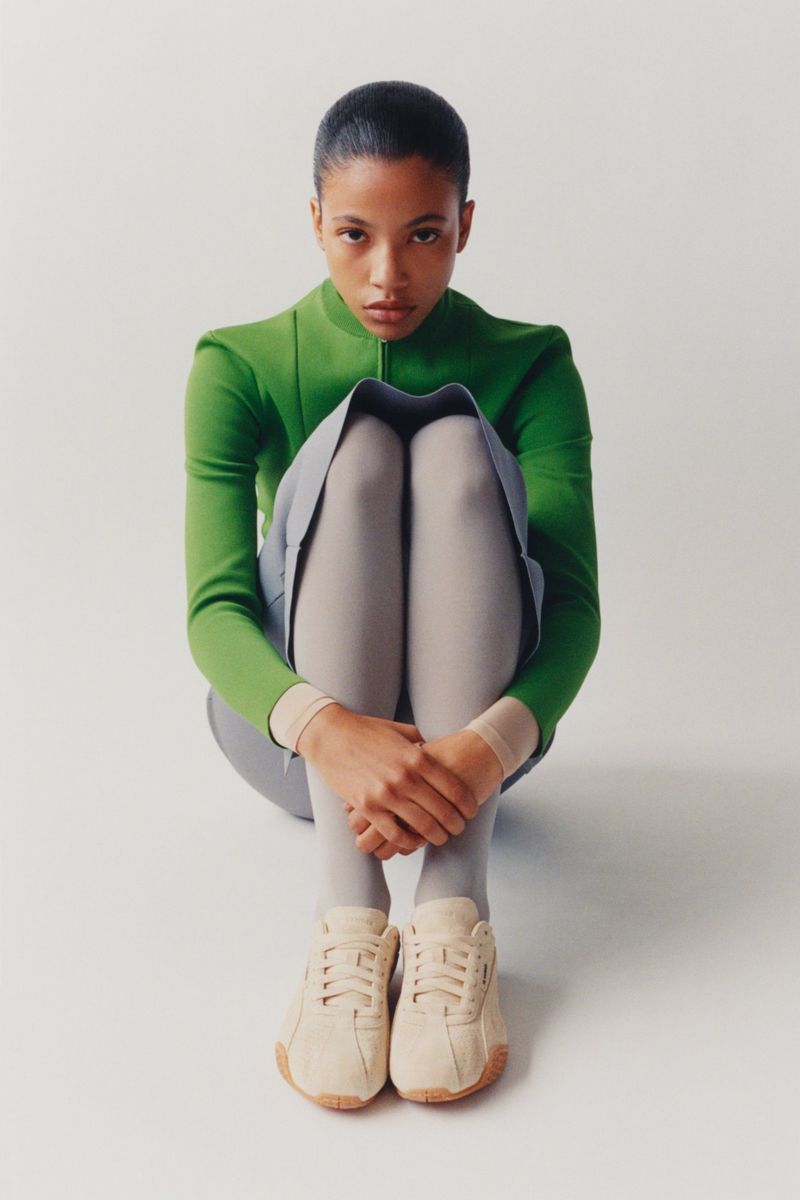 Model in a green top and grey skirt sitting on the floor wearing beige sneakers against a seamless white background