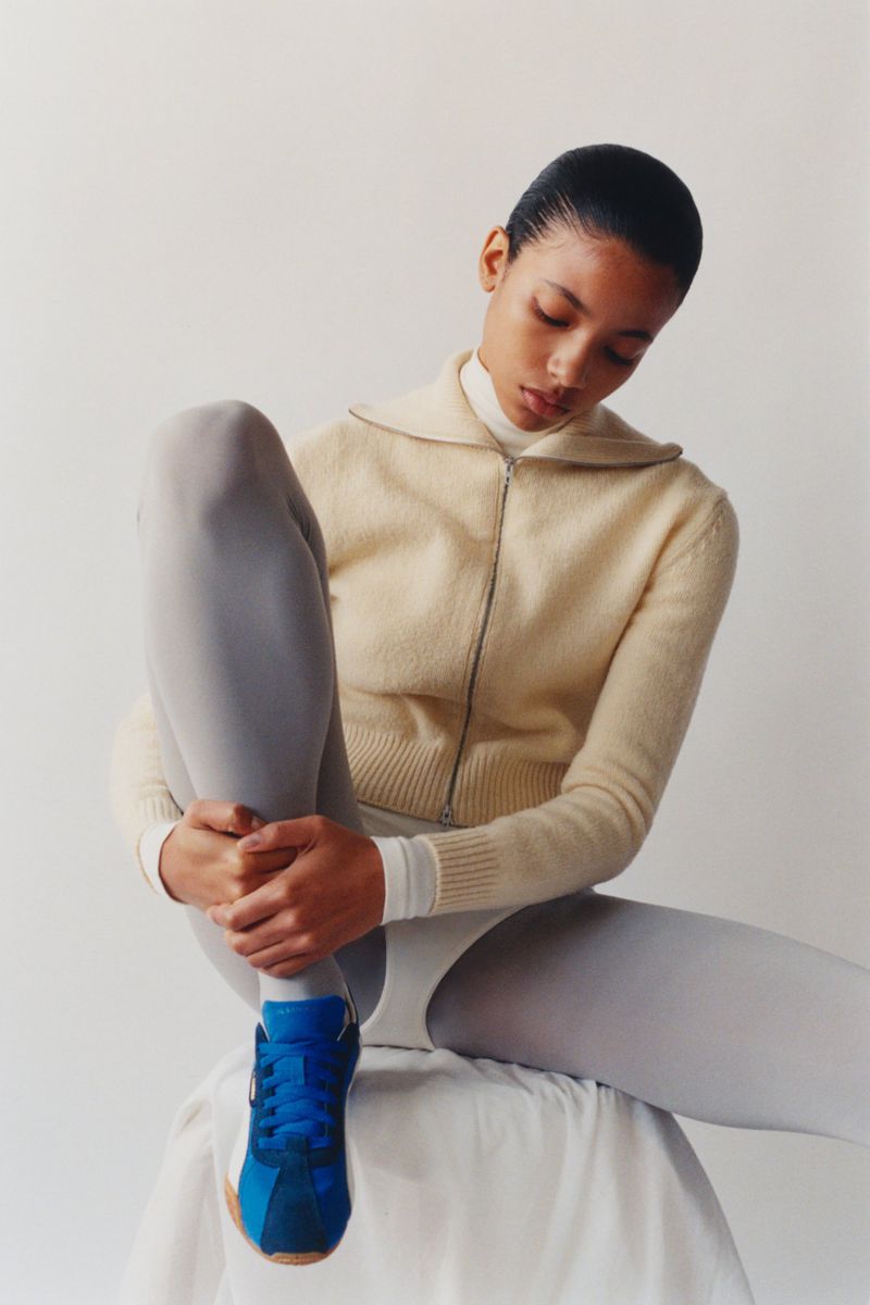 Young woman in cream sweater, grey leggings and blue sneakers poses seated on a white stool against a plain white background.