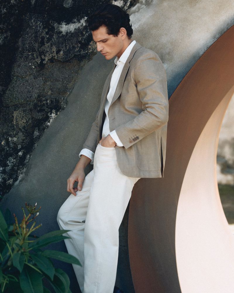 Model Pau Ramis leaning against an organic curved concrete structure wearing a tailored beige blazer and white trousers.