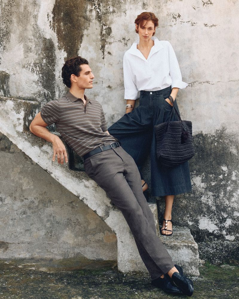 Man leaning on a concrete staircase wearing a striped polo shirt next to a woman standing in a white blouse and dark trousers