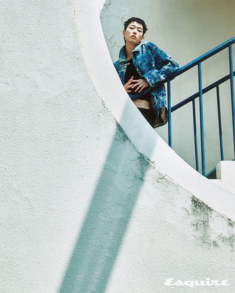 Young man with short hair, wearing a denim jacket, poses on a stairway with white walls and blue railings in an outdoor setting.