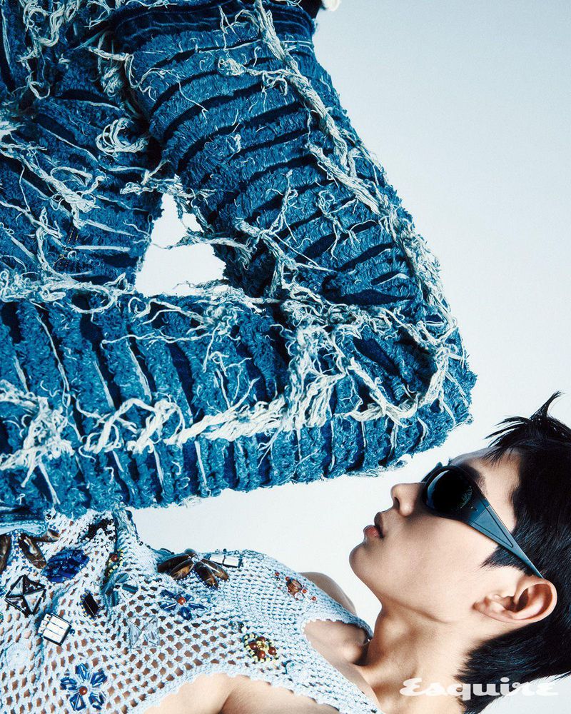 Fashion editorial portrait: Man with dark hair poses in a blue textured shirt and sunglasses against a white background.