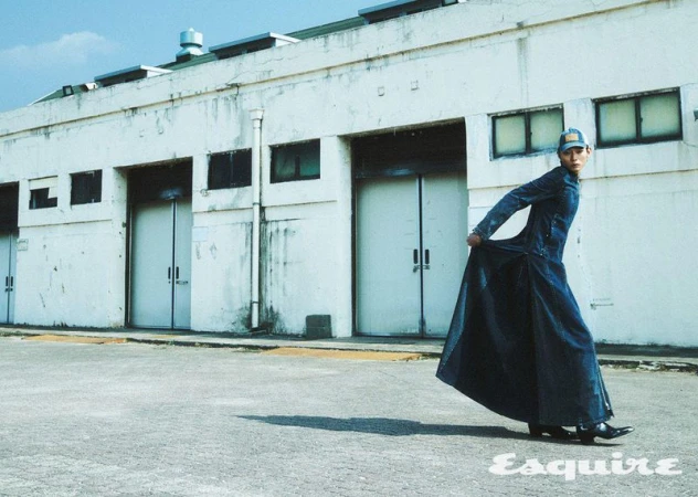 An adult male model in a long denim coat and baseball cap poses in front of a white industrial building. Esquire logo at the lower right.
