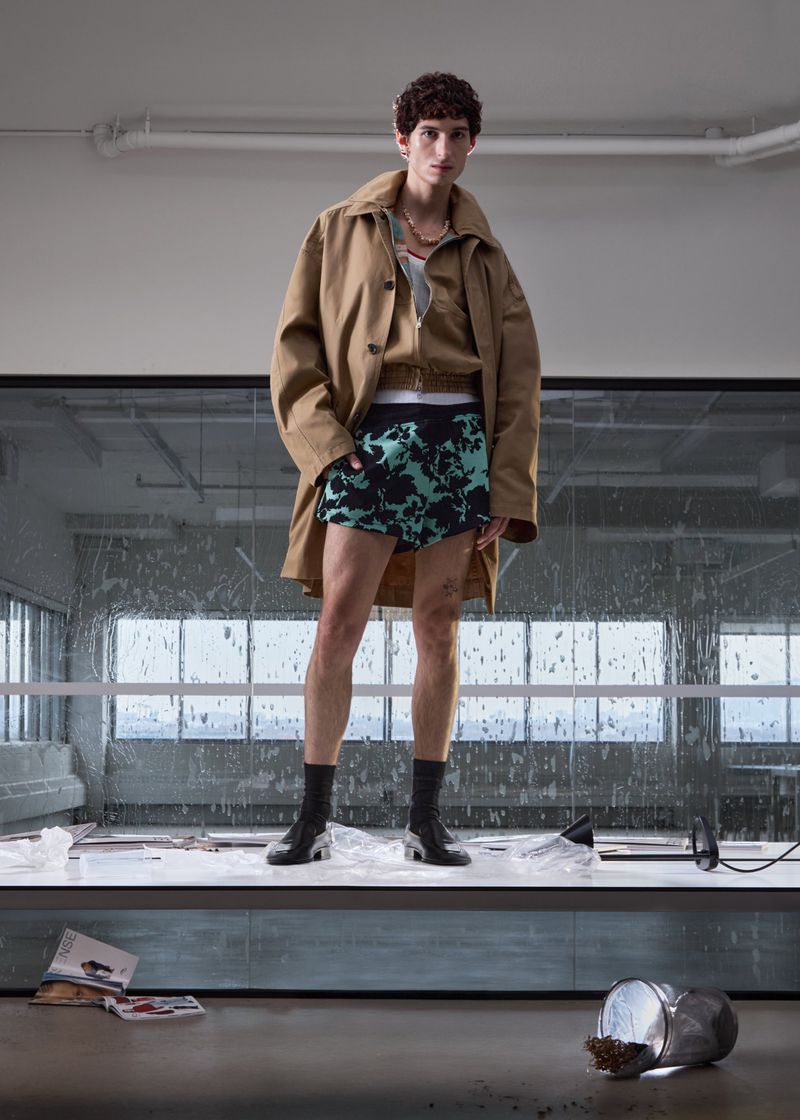 Model wearing a tan jacket and patterned shorts standing on a white surface inside an industrial space by Geray Mena for SSENSE.