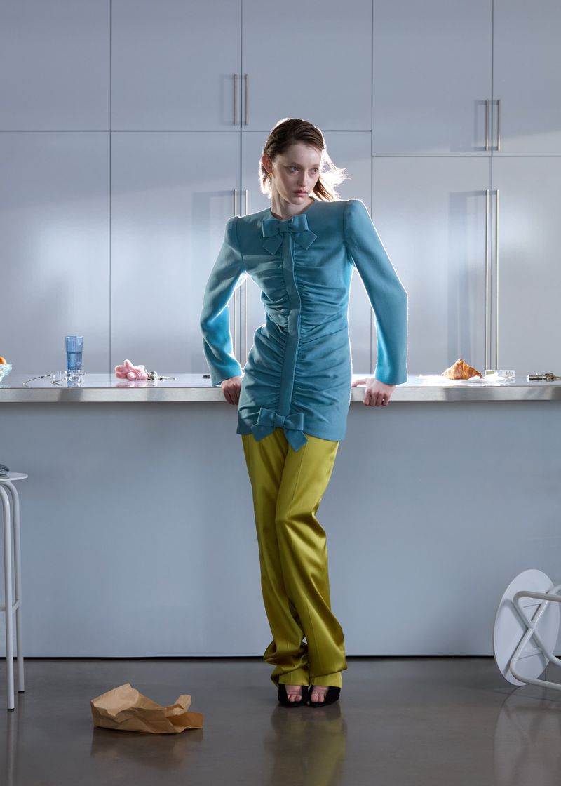 A fashion model poses in a blue dress with olive green pants, leaning against a kitchen counter in a modern, minimalist kitchen setting.