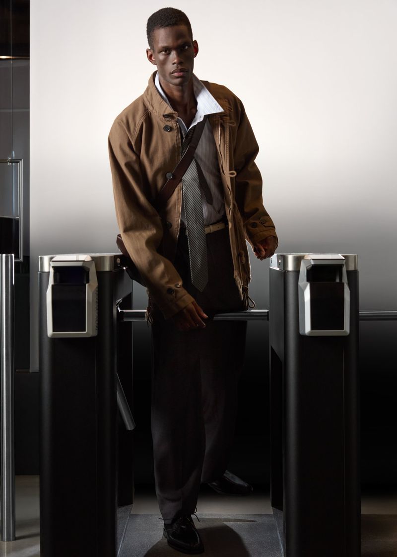 Man wearing a brown jacket and tie posing between turnstiles in an SSENSE campaign shot by Geray Mena.