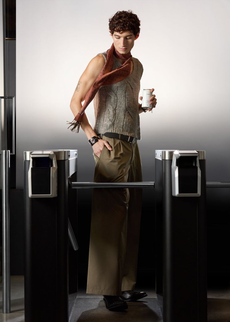 Man wearing a grey knit vest and scarf standing at turnstiles for SSENSE S/S 26 campaign photographed by Geray Mena.