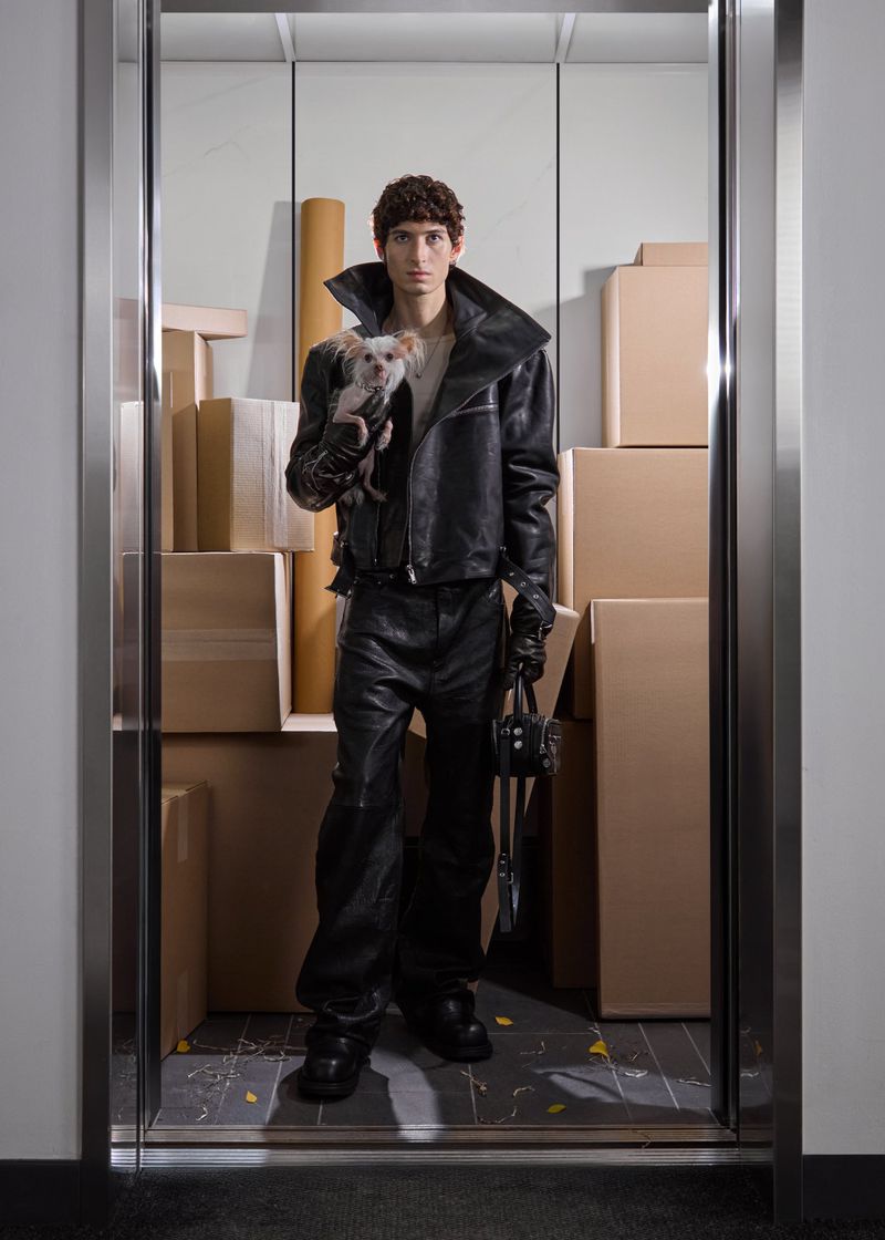 Model wearing black leather jacket and pants holding a small dog inside an elevator filled with cardboard boxes
