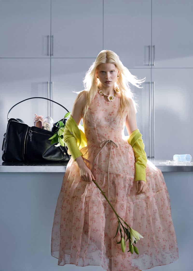 Blonde model in a floral pink dress and yellow sweater holding a white lily in a modern kitchen with a large black bag.