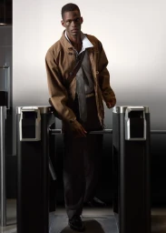 Man wearing a brown jacket and tie posing between turnstiles in an SSENSE campaign shot by Geray Mena.