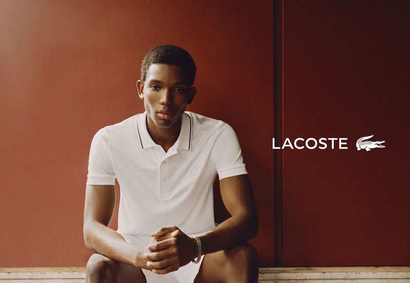 Model Ibra Niakate seated wearing a white polo shirt against a terracotta wall in a Lacoste campaign.