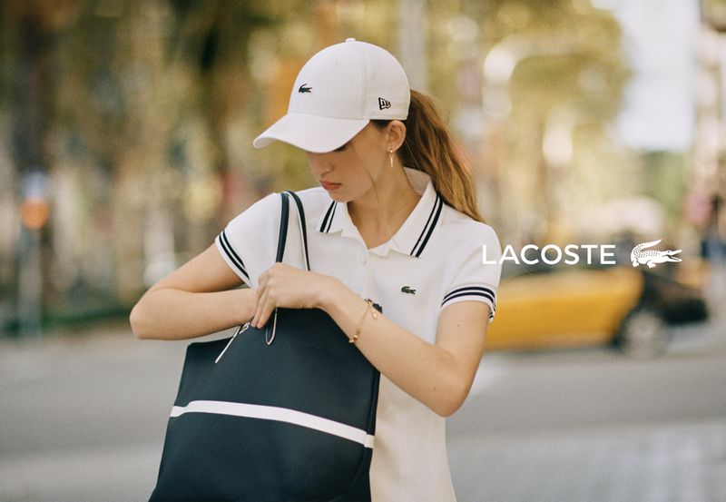 Model Elise Marie wearing a white Lacoste cap and polo shirt searching through a navy blue tote bag on a city street.