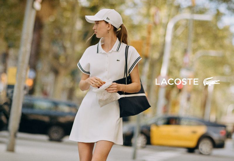 Model in white polo dress and baseball cap holding a tote bag on a city street for Lacoste Tennis Attitude Spring 2026.