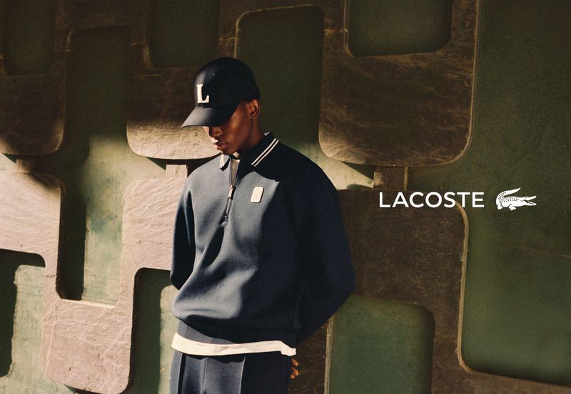 A man wearing a dark polo shirt and baseball cap leaning against a patterned wall for Lacoste.