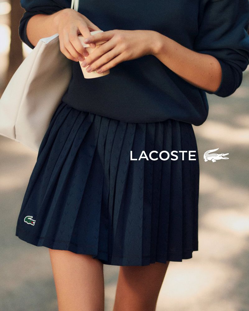 Model wearing a navy pleated skirt and sweatshirt holding a cup and white bag for Lacoste Tennis Attitude Spring 2026