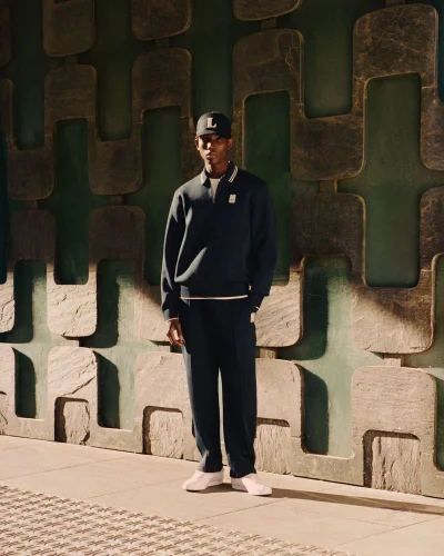 Model Ibra Niakate posing in a dark navy zip polo sweatshirt, matching trousers, and a baseball cap against a geometric textured wall.