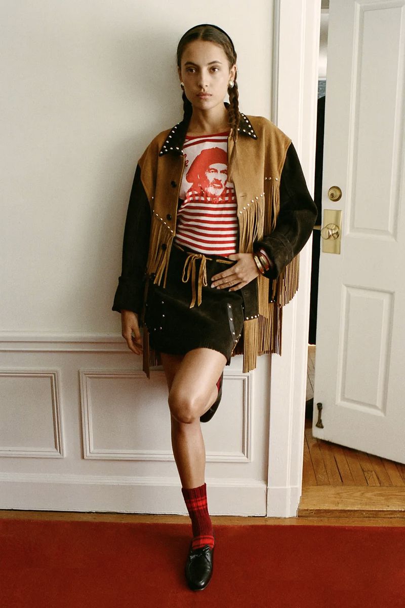 Alyssa Morelle poses in a fringed suede jacket, red striped graphic shirt, and black mini skirt by an open white door.